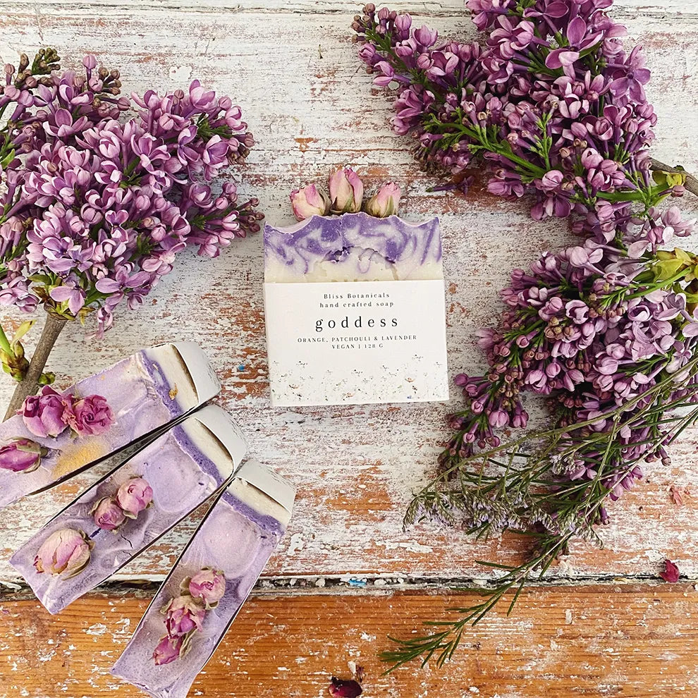 Bar of goddess soap  surrounded by purple flowers on a wooden surface