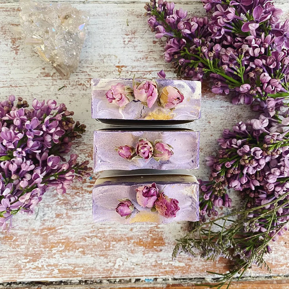Three bars of goddess soap with floral embedded ingredients on a wooden surface with lavender flowers.
